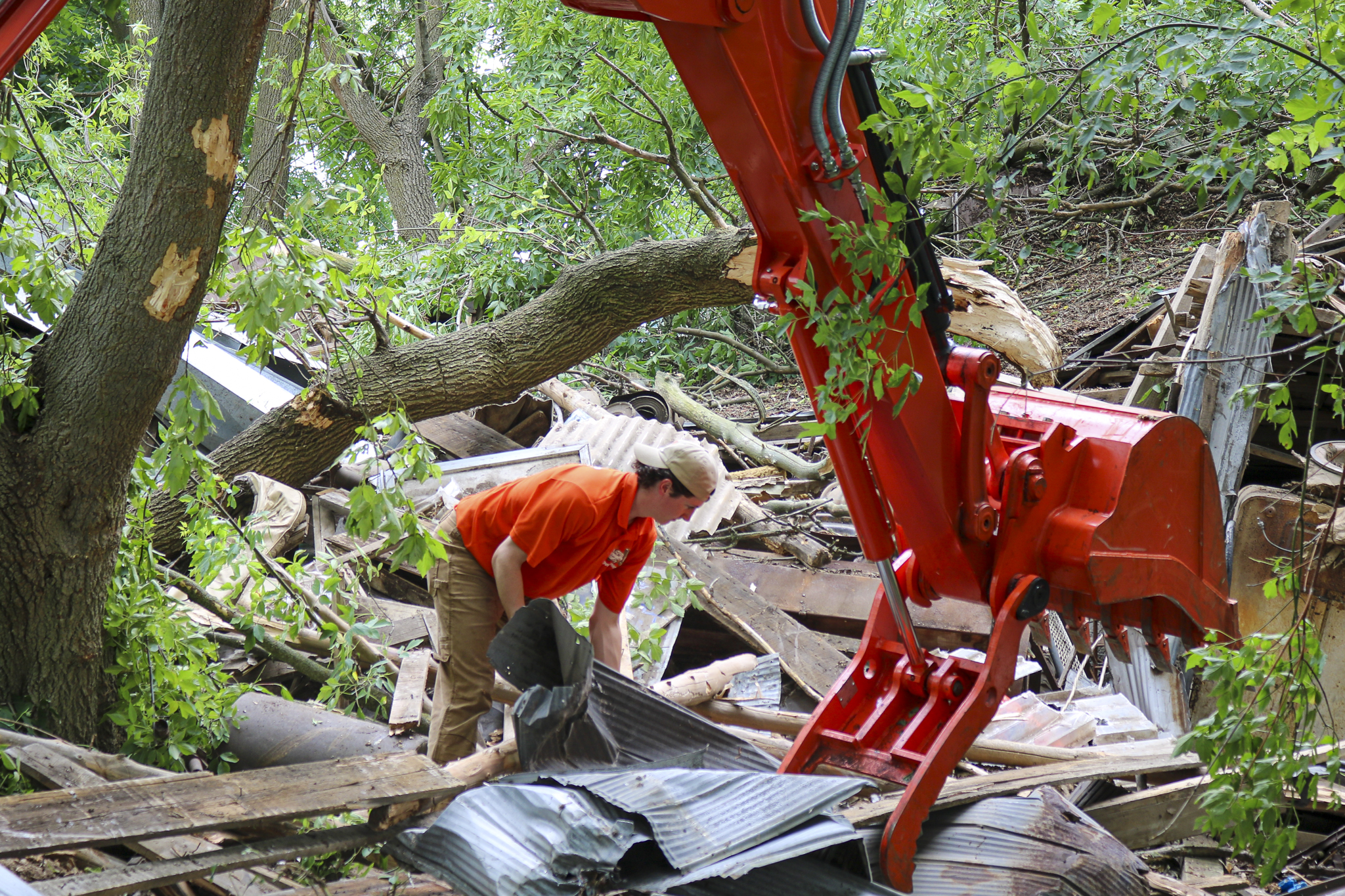 A worker in an orange shirt is actively clearing debris with an excavator in a cluttered area surrounded by trees.